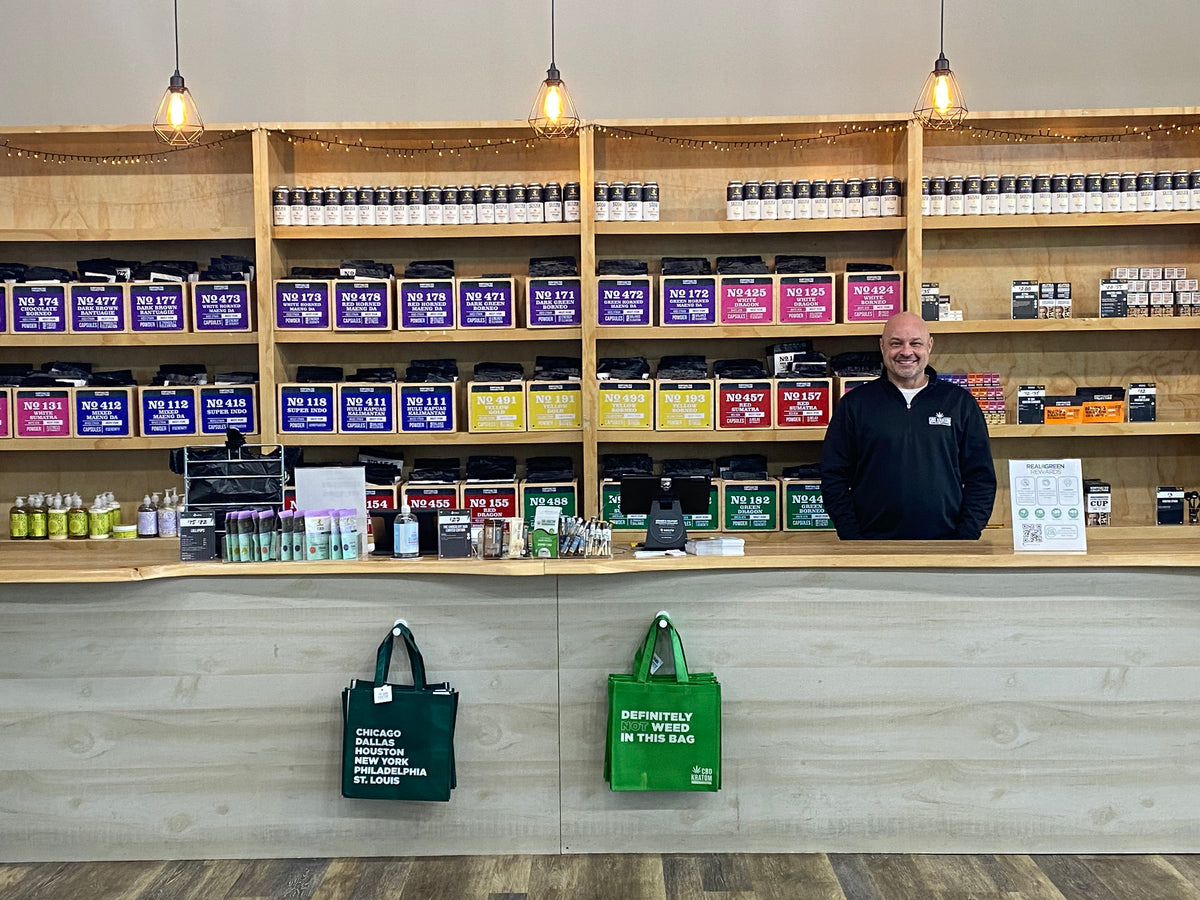 A CBD Kratom team member in a branded quarter-zip smiling behind the counter in front of the kratom wall at the St. Peters store near St. Louis.